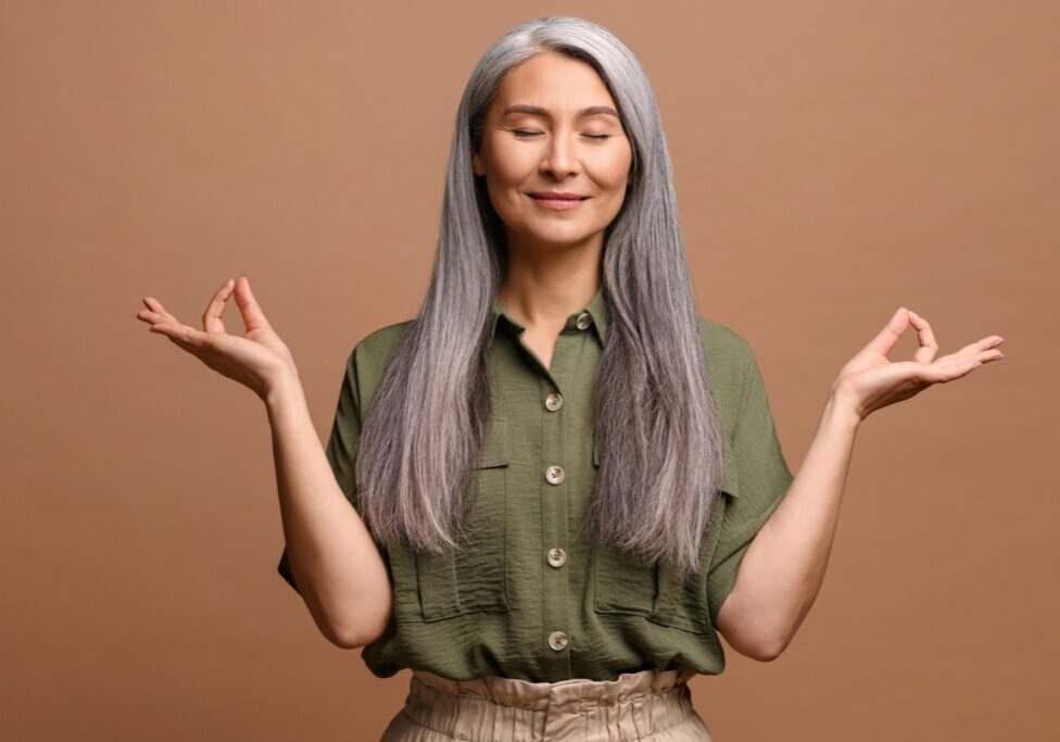Meditation concept. Calm and satisfied mature woman holds her hands in zen gesture, keeps eyes closed, feels appeasement and pacification, practicing yoga, studio shot isolated on brown